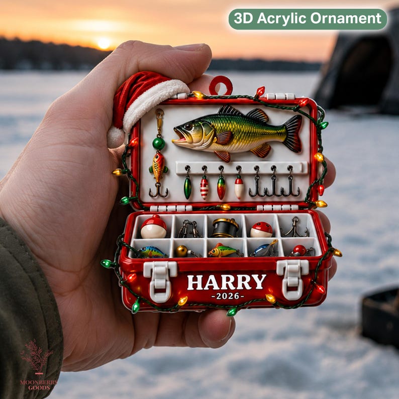 May include: A 3D acrylic ornament shaped like a red fishing tackle box with a Santa hat on top. The box contains a fish figure, various lures, and hooks. The front is printed with the name HARRY and the year 2026, decorated with string lights.