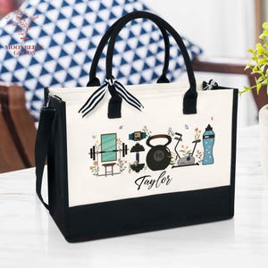 May include: A black and white tote bag with black handles and a shoulder strap. The bag features a design of fitness equipment, flowers, and the name "Taylor". A black and white striped bow is tied to the handles.