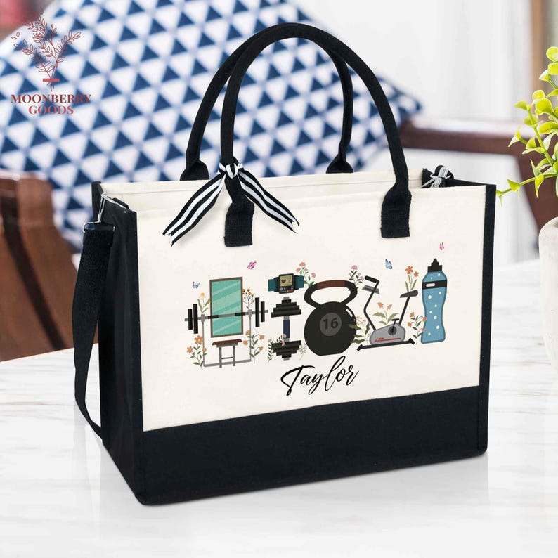 May include: A black and white tote bag with black handles and a shoulder strap. The bag features a design of fitness equipment, flowers, and the name "Taylor". A black and white striped bow is tied to the handles.