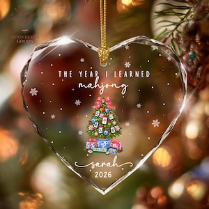 May include: A heart-shaped clear acrylic ornament featuring a printed Christmas tree decorated with mahjong tiles, a blue and white patterned pot base, and gift boxes. The text reads THE YEAR I LEARNED mahjong, sarah, and 2026, with white snowflakes.