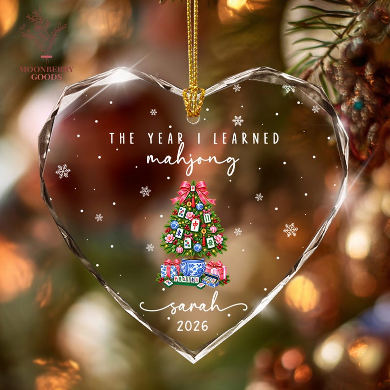 May include: A heart-shaped clear acrylic ornament featuring a printed Christmas tree decorated with mahjong tiles, a blue and white patterned pot base, and gift boxes. The text reads THE YEAR I LEARNED mahjong, sarah, and 2026, with white snowflakes.