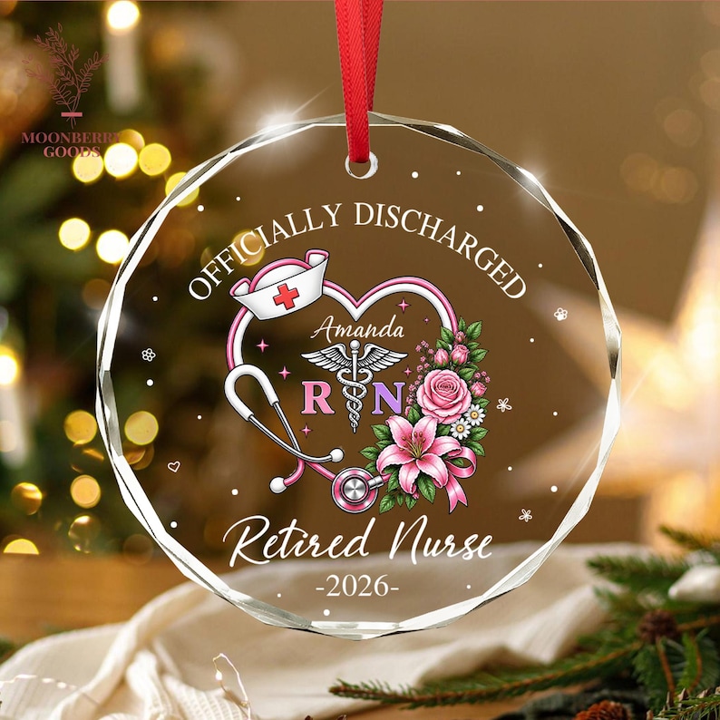 May include: A circular clear glass ornament with a red ribbon hanger. The design features a pink heart, a stethoscope, a nurse cap, a caduceus symbol, and pink flowers. Text reads: OFFICIALLY DISCHARGED, Amanda, RN, Retired Nurse, and 2026.