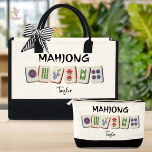 May include: A white tote bag and matching zippered pouch with a black base. Both feature the word "MAHJONG" above colorful Mahjong tiles. The tote has black handles and a striped bow. The name "Taylor" is printed below the tiles.