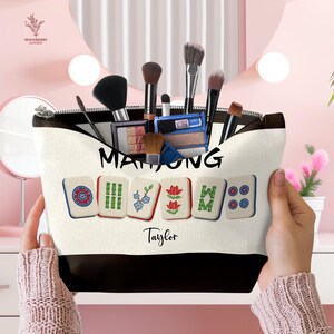May include: A cream-colored makeup bag with a black zipper and bottom, filled with makeup brushes and products. The bag features the word "MAHJONG" and Mahjong tile graphics. The name "Taylor" is printed below the tiles.