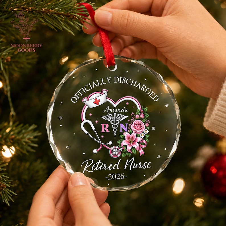 May include: A round, clear, faceted glass ornament hanging from a red ribbon. The design features a pink heart, a stethoscope, a nurse cap, and flowers. Text reads: Officially Discharged, Amanda, RN, Retired Nurse, 2026. The ornament has a reflective surface.