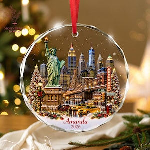 May include: A round, faceted glass ornament featuring a New York City skyline scene with the Statue of Liberty, yellow taxis, and a Broadway sign. The design includes decorated Christmas trees, snow, and the custom text Amanda 2026 in red script.