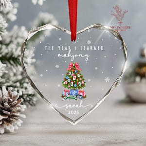May include: A heart-shaped clear acrylic ornament with a red ribbon hanger. The text reads THE YEAR I LEARNED mahjong, sarah, and 2026. A graphic of a Christmas tree decorated with mahjong tiles, flowers, and ornaments sits above wrapped gifts.