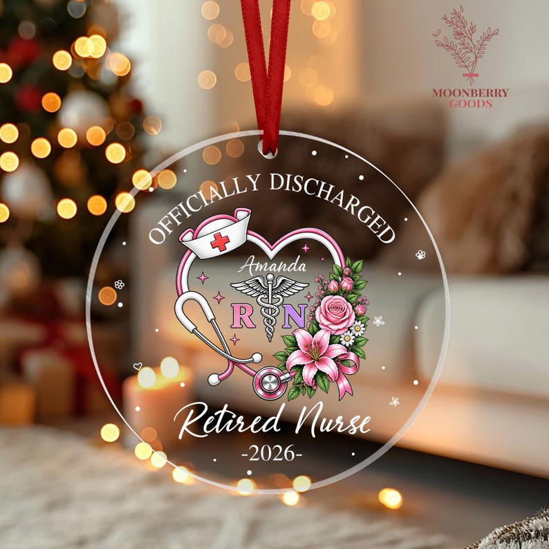 May include: A circular clear acrylic ornament with a red ribbon hanger. The design features a pink stethoscope forming a heart shape, a nurse cap, a caduceus symbol, pink flowers, and the text: OFFICIALLY DISCHARGED, Amanda, RN, Retired Nurse, 2026.