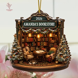 May include: A decorative hanging ornament shaped like a bookstore interior with a sign reading AMANDA'S BOOKSTORE 2026. It features two armchairs, a table with an open book, a sleeping cat, and two Christmas trees with lights and snow accents.