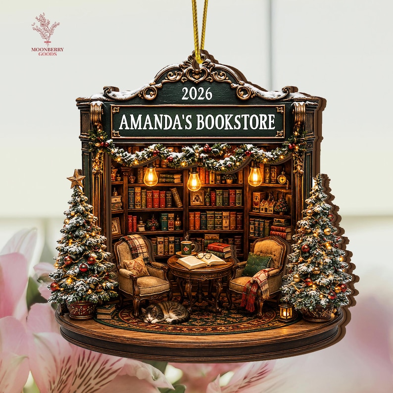 May include: A decorative hanging ornament shaped like a bookstore interior with a sign reading AMANDA'S BOOKSTORE 2026. It features two armchairs, a table with an open book, a sleeping cat, and two Christmas trees with lights and snow accents.