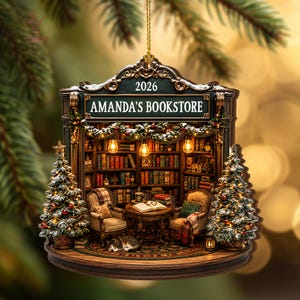 May include: A hanging ornament shaped like a bookstore interior with the text AMANDA'S BOOKSTORE and 2026. It features two armchairs, a round table with an open book, a sleeping cat, bookshelves, and two decorated Christmas trees on the sides.
