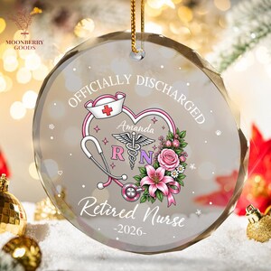 May include: A round, clear glass ornament featuring the text "OFFICIALLY DISCHARGED" and "Retired Nurse -2026-". The design includes a pink stethoscope forming a heart, a nurse cap with a red cross, a caduceus symbol, and pink floral accents.