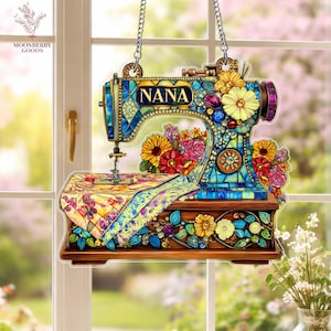 May include: A decorative sewing machine ornament with the word "NANA" on the front. The ornament is adorned with colorful flowers and jewels. It hangs from a silver chain and is set against a window.