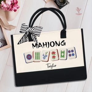 May include: Cream-colored tote bag with black trim and handles. The bag features the word "MAHJONG" in black letters above illustrated Mahjong tiles. A black and white striped bow is attached to the handle. The name "Taylor" is printed below the tiles.