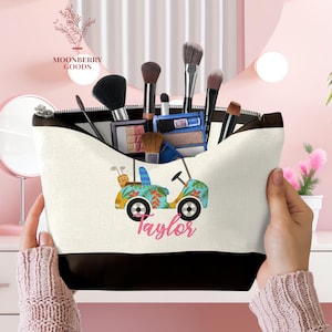 Op de afbeelding: Een witte en zwarte make-uptas met een golfkar-ontwerp in blauw, geel en groen, met de naam "Taylor" in roze script. De tas is gevuld met make-upborstels en -producten.