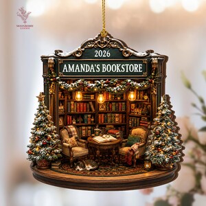 May include: A decorative hanging ornament shaped like a bookstore interior with the text AMANDA'S BOOKSTORE and 2026. It features two armchairs, a table with an open book, a sleeping cat, bookshelves, and two snow-dusted Christmas trees with lights.