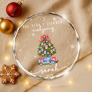 May include: A round, clear acrylic Christmas ornament featuring the text "THE YEAR I LEARNED mahjong." and "sarah 2026". The design includes a green Christmas tree decorated with mahjong tiles, red bows, and ornaments, with gifts at the base.