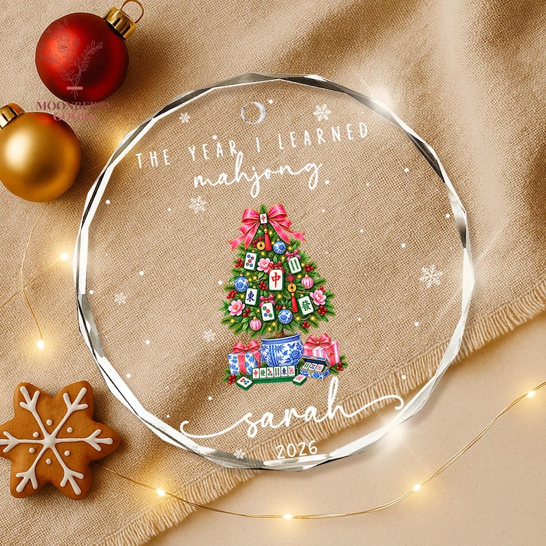 May include: A round, clear acrylic Christmas ornament featuring the text "THE YEAR I LEARNED mahjong." and "sarah 2026". The design includes a green Christmas tree decorated with mahjong tiles, red bows, and ornaments, with gifts at the base.