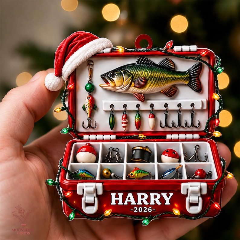 May include: A red tackle box ornament featuring a Santa hat, a bass fish, and various fishing lures. The interior compartments hold hooks and bobbers. The exterior is decorated with string lights and the white text HARRY 2026 printed on the front.