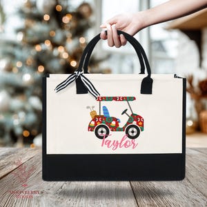 Op de afbeelding: Een canvas draagtas met zwarte handvatten en een zwarte basis. De tas heeft een kleurrijk golfkarretje ontwerp met de naam "Taylor" in roze. Een zwart-wit gestreept lint is aan het handvat geknoopt.