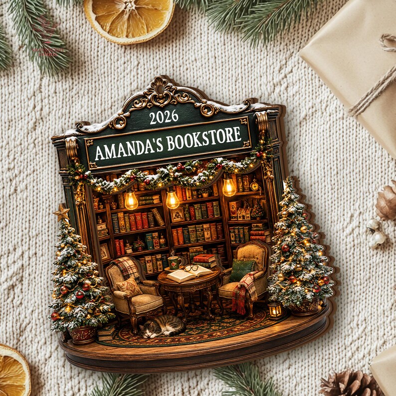 May include: A decorative ornament shaped like a bookstore storefront with the text 2026 AMANDA'S BOOKSTORE. It features shelves filled with books, two armchairs, a small table with an open book, a sleeping cat, and two lit Christmas trees.