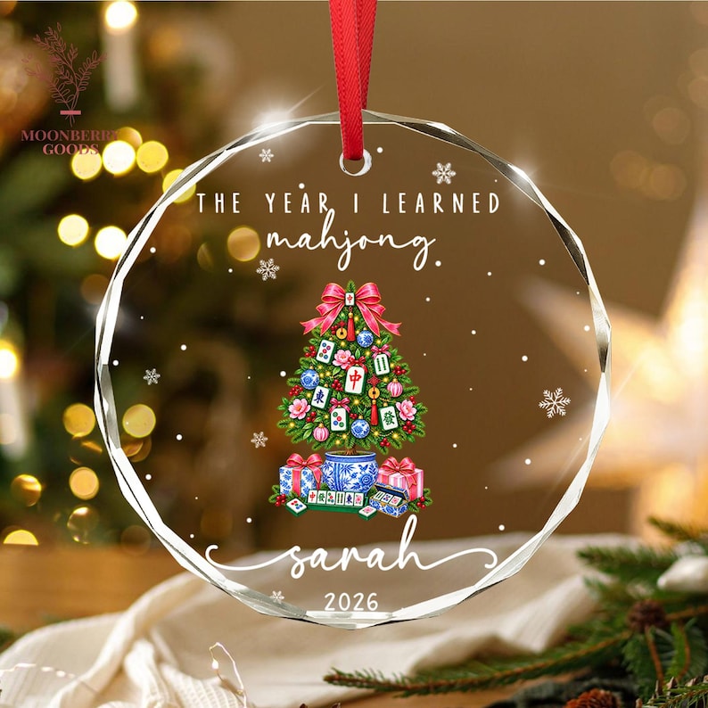 May include: A round, faceted clear glass ornament hanging from a red ribbon. It features the text THE YEAR I LEARNED mahjong. above a green Christmas tree decorated with mahjong tiles, followed by the name sarah and the year 2026 below.