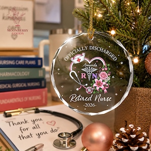 May include: A round, clear, faceted glass ornament featuring a printed design with a stethoscope, a nurse cap, pink flowers, and the text "OFFICIALLY DISCHARGED", "Amanda", "RN", "Retired Nurse", and "2026". It hangs from a gold cord.