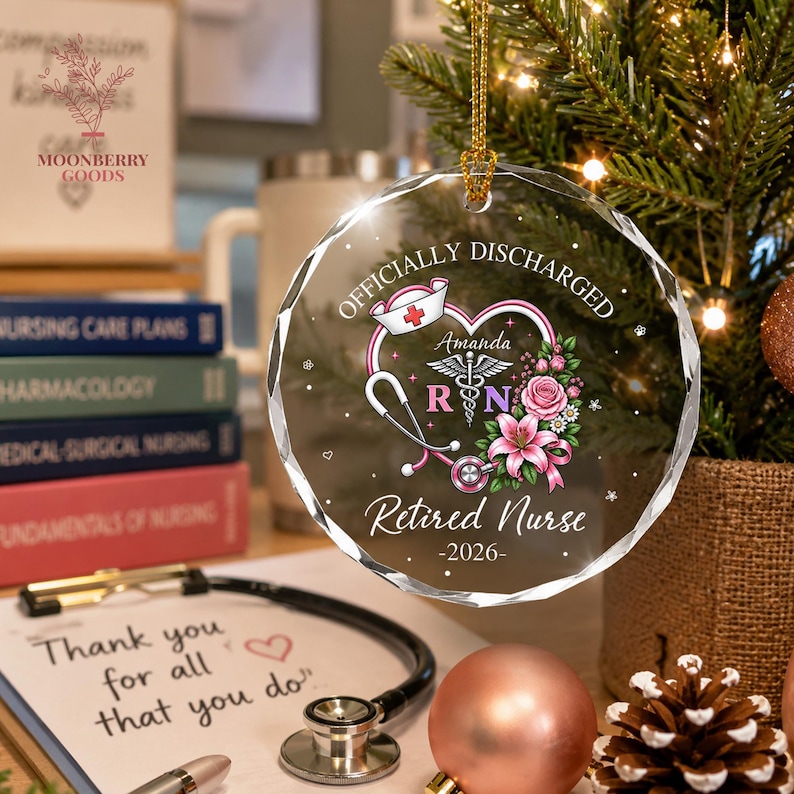 May include: A round, clear, faceted glass ornament featuring a printed design with a stethoscope, a nurse cap, pink flowers, and the text "OFFICIALLY DISCHARGED", "Amanda", "RN", "Retired Nurse", and "2026". It hangs from a gold cord.