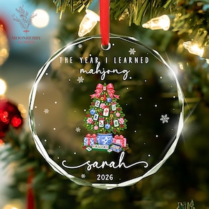 May include: A clear, circular, faceted glass Christmas ornament hanging from a red ribbon. It features a green Christmas tree decorated with mahjong tiles, blue and pink ornaments, and a red bow. Text reads: THE YEAR I LEARNED mahjong. sarah 2026.