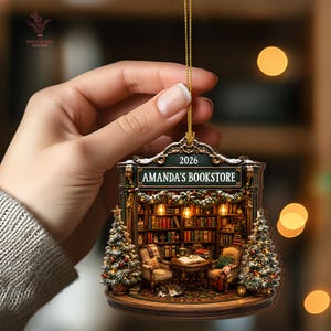 May include: A decorative hanging ornament featuring a detailed bookstore interior with shelves of books, two armchairs, a small table, and a cat. The sign reads AMANDA'S BOOKSTORE and 2026. Two Christmas trees flank the entrance, all suspended by a gold cord.