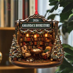 May include: A decorative hanging ornament shaped like a bookstore interior with the text AMANDA'S BOOKSTORE and 2026. It features two armchairs, a table with an open book, a sleeping cat, bookshelves, and two snow-dusted Christmas trees with lights.