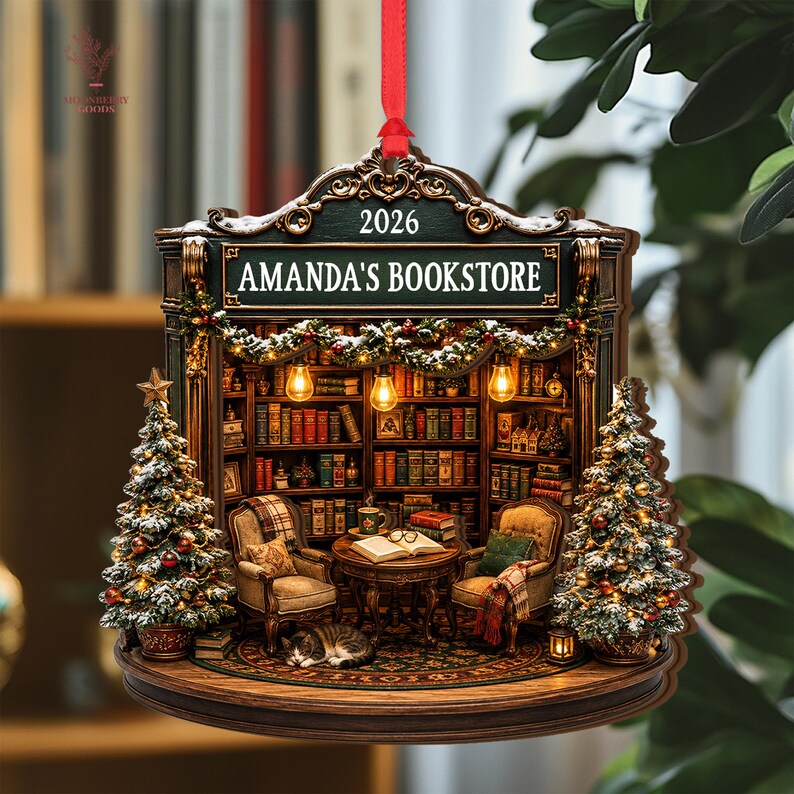 May include: A decorative hanging ornament shaped like a bookstore interior with the text AMANDA'S BOOKSTORE and 2026. It features two armchairs, a table with an open book, a sleeping cat, bookshelves, and two snow-dusted Christmas trees with lights.