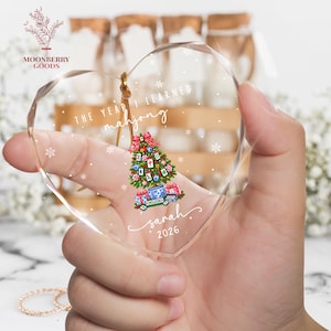May include: A heart-shaped clear acrylic ornament featuring a printed graphic of a Christmas tree decorated with mahjong tiles, a red bow, and gifts. The text reads THE YEAR I LEARNED mahjong, sarah, and 2026, surrounded by white snowflake accents.