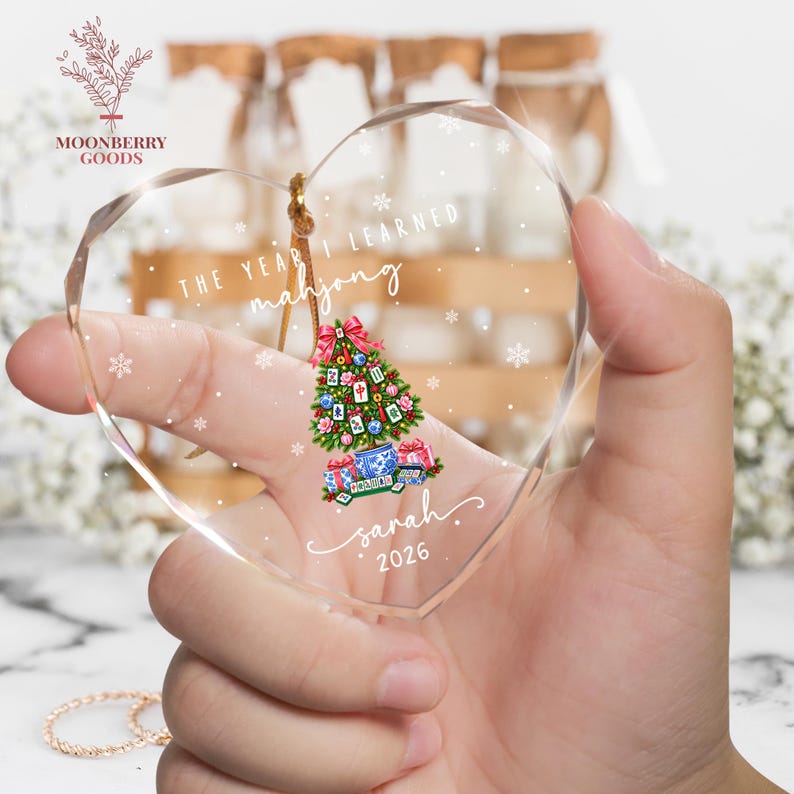 May include: A heart-shaped clear acrylic ornament featuring a printed graphic of a Christmas tree decorated with mahjong tiles, a red bow, and gifts. The text reads THE YEAR I LEARNED mahjong, sarah, and 2026, surrounded by white snowflake accents.