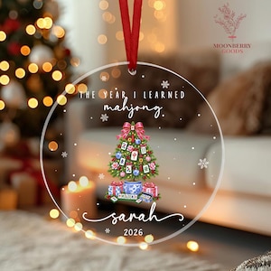 May include: A clear circular acrylic ornament with a red ribbon hanger. It features the text THE YEAR I LEARNED mahjong. and sarah 2026. The center graphic shows a Christmas tree decorated with mahjong tiles, ornaments, and a blue and white pot base.