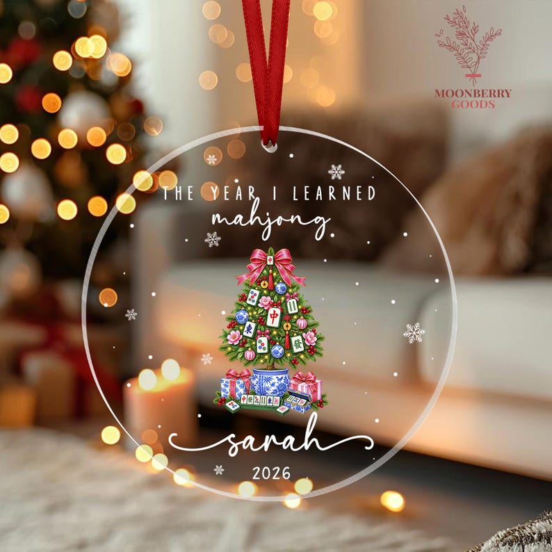 May include: A clear circular acrylic ornament with a red ribbon hanger. It features the text THE YEAR I LEARNED mahjong. and sarah 2026. The center graphic shows a Christmas tree decorated with mahjong tiles, ornaments, and a blue and white pot base.