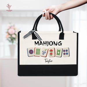 May include: A canvas tote bag with black handles and trim, featuring the word "MAHJONG" in black letters. Below the word are six Mahjong tiles. The name "Taylor" is printed in black script below the tiles. The bag is held by a person.