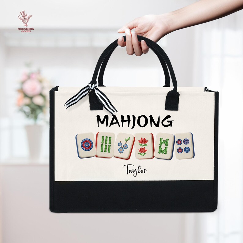 May include: A canvas tote bag with black handles and trim, featuring the word "MAHJONG" in black letters. Below the word are six Mahjong tiles. The name "Taylor" is printed in black script below the tiles. The bag is held by a person.