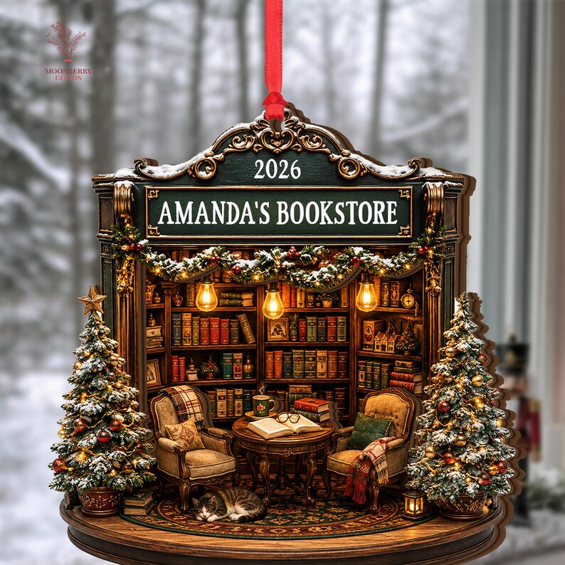 May include: A decorative hanging ornament shaped like a bookstore interior with the text 2026 and AMANDA'S BOOKSTORE. The scene features two armchairs, a table with an open book, a cat, and two Christmas trees decorated with lights and ornaments.
