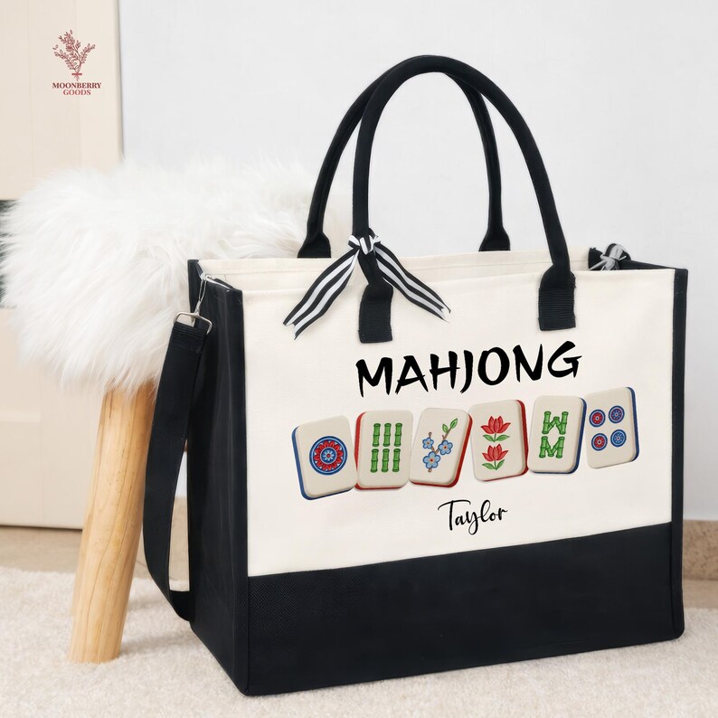 May include: A canvas tote bag with black handles and a black bottom panel. The bag features the word "MAHJONG" in black, along with colorful Mahjong tile graphics. The name "Taylor" is printed below the tiles.