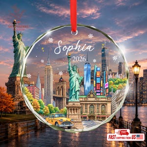 May include: A circular glass ornament with a red ribbon featuring a New York City skyline collage, the Statue of Liberty, a yellow taxi, and the text Sophia 2026. The design includes snowflakes and stars against a sunset sky background.