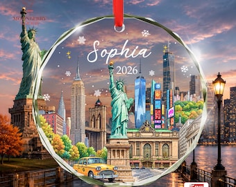 May include: A circular glass ornament with a red ribbon featuring a New York City skyline collage, the Statue of Liberty, a yellow taxi, and the text Sophia 2026. The design includes snowflakes and stars against a sunset sky background.