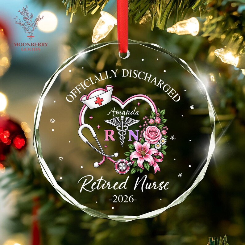 May include: A circular clear glass ornament with a red ribbon, featuring the text "OFFICIALLY DISCHARGED" and "Retired Nurse -2026-". The design includes a nurse cap, a stethoscope forming a heart, a caduceus symbol, and pink floral accents.