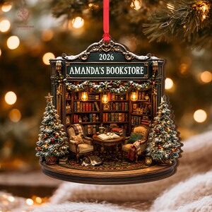 May include: A decorative hanging ornament featuring a detailed bookstore interior with shelves of books, two armchairs, a small table, a sleeping cat, and two Christmas trees. The sign reads AMANDA'S BOOKSTORE and 2026. It hangs from a red ribbon.
