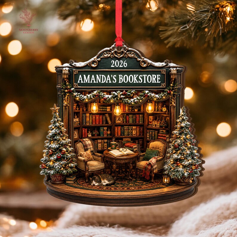 May include: A decorative hanging ornament featuring a detailed bookstore interior with shelves of books, two armchairs, a small table, a sleeping cat, and two Christmas trees. The sign reads AMANDA'S BOOKSTORE and 2026. It hangs from a red ribbon.