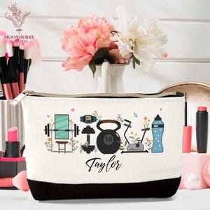 May include: A white canvas cosmetic bag with a black bottom and gold zipper. The bag features a fitness-themed design with a mirror, dumbbells, kettlebell, exercise bike, and water bottle. The name "Taylor" is written on the bag.