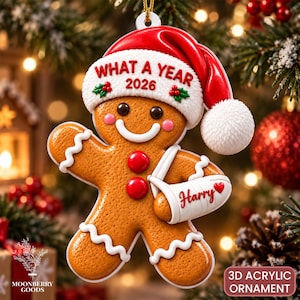 May include: A gingerbread person Christmas ornament wearing a red Santa hat with the text WHAT A YEAR 2026 and holly accents. The figure has two red buttons and a white arm sling with the name Harry and a small red heart printed on it.