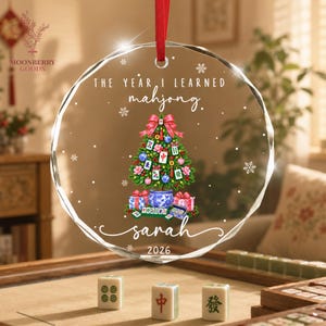 May include: A round, clear acrylic ornament with a red ribbon hanger. It features the text "THE YEAR I LEARNED mahjong." above a Christmas tree decorated with mahjong tiles, ornaments, and a bow. The name "sarah" and "2026" are printed below.