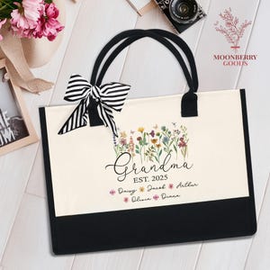 May include: A white canvas tote bag with black handles and trim. The bag features a floral design with the text "Grandma Est. 2025" and a list of names: Daisy, Jacob, Arthur, Olivia, Diana.