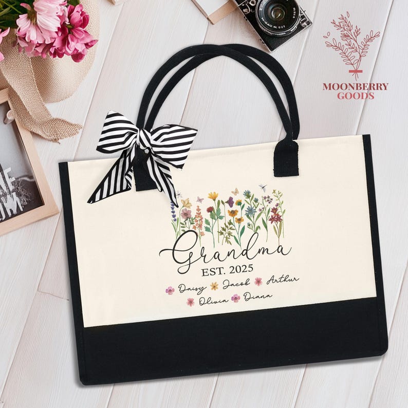 May include: A white canvas tote bag with black handles and trim. The bag features a floral design with the text "Grandma Est. 2025" and a list of names: Daisy, Jacob, Arthur, Olivia, Diana.